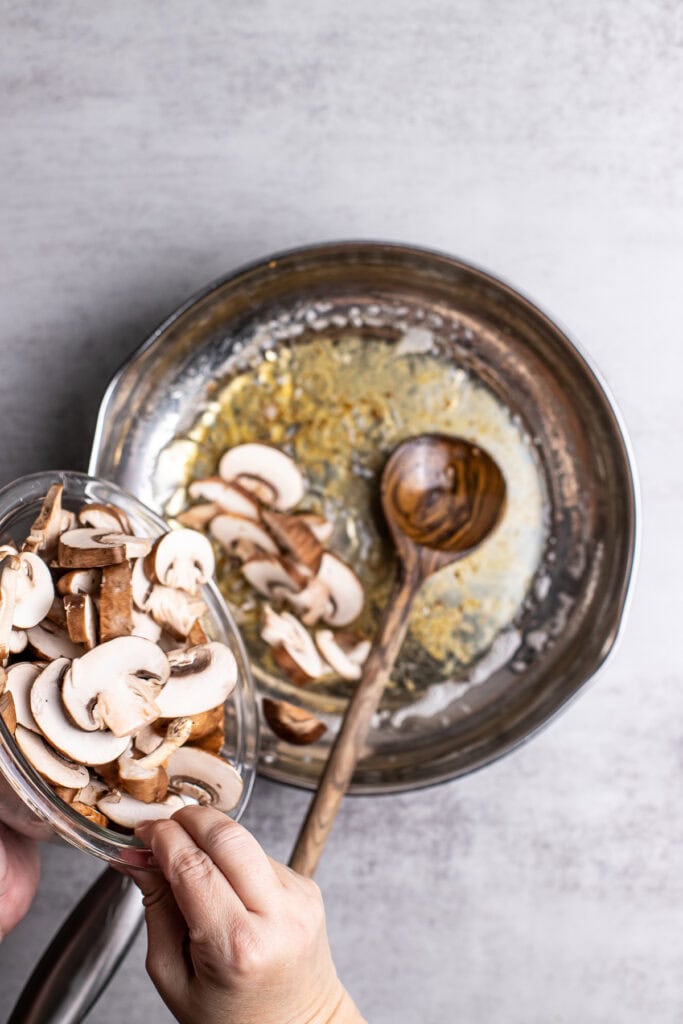 Fresh sliced mushrooms being added to sautéed mushrooms and garlic in a skillet, perfect for mushroom recipes and cooking tips.