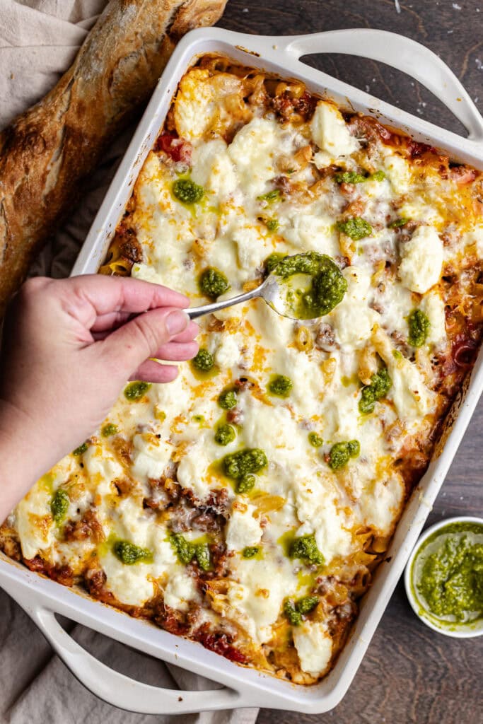 Creamy no-boil penne pasta casserole with ground beef & cheese and pesto, served in a white baking dish with crusty bread on the side. Perfect comfort food recipe for family dinners or casual gatherings.