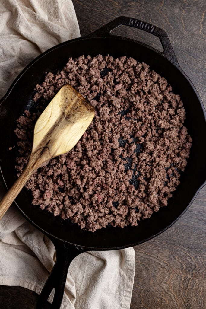 Cooked ground beef in a black skillet with a wooden spatula in a rustic kitchen setting.