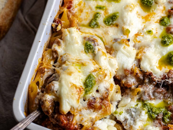 Creamy baked lasagna with melted cheese, seasoned meat, and green pesto topping in a white casserole dish, served with toasted garlic bread for a hearty Italian meal.