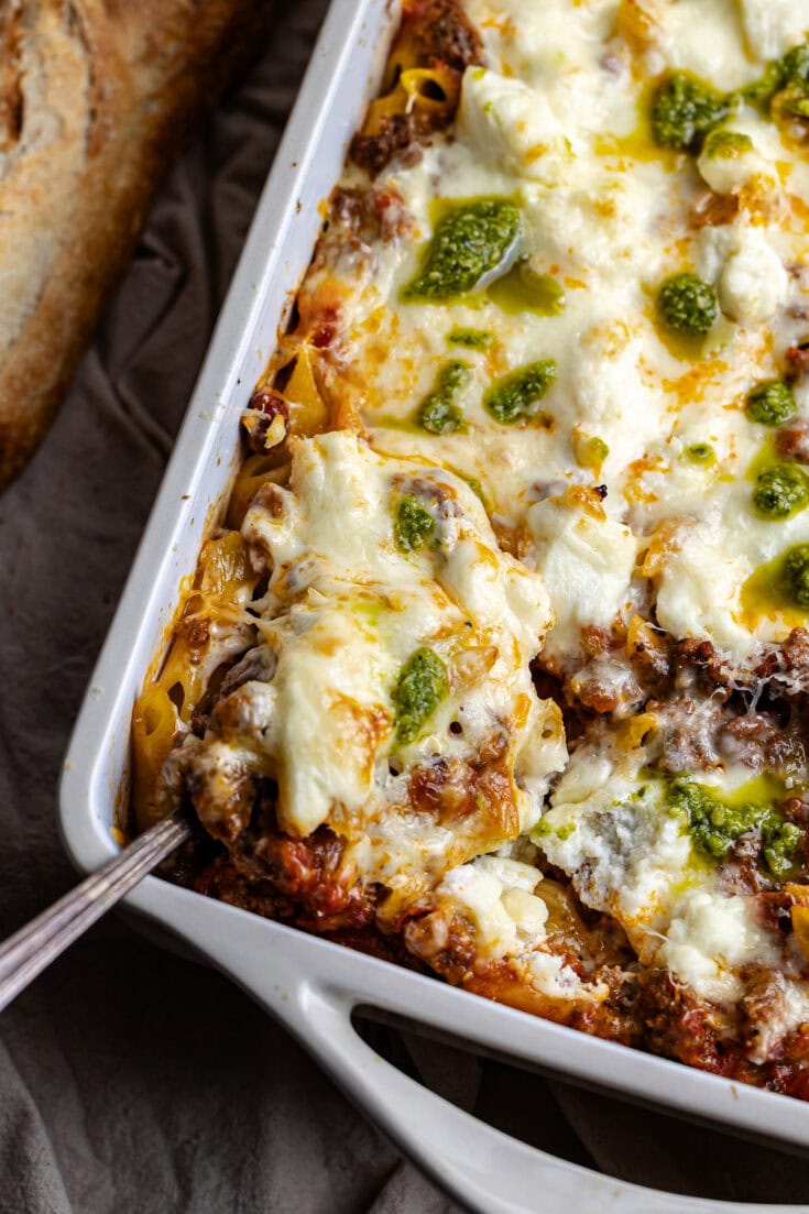 Creamy baked lasagna with melted cheese, seasoned meat, and green pesto topping in a white casserole dish, served with toasted garlic bread for a hearty Italian meal.