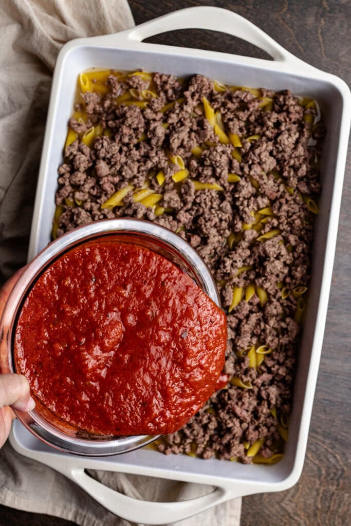 Ground beef and tomato sauce layered over cooked pasta in a baking dish for a homemade lasagna recipe.
