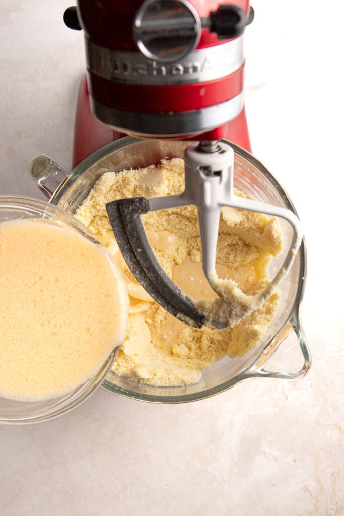 Flour and eggs being mixed in a stand mixer for baking, with a pour of beaten eggs in a glass bowl, ideal for baking recipes at Stuck On Sweet.