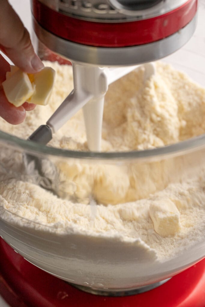 Butter being added to a mixing bowl with flour and a stand mixer for baking cookies or cakes | butter in a stand mixer for baking sweet treats | mixing butter and flour for baking cookies or cake batter at Stuck On Sweet | close-up of butter addition to dry ingredients in a stand mixer, preparing for baking delicious desserts | relevance: relevant for baking, baking ingredients, cookie, cake, dessert, kitchen, sweet treats.