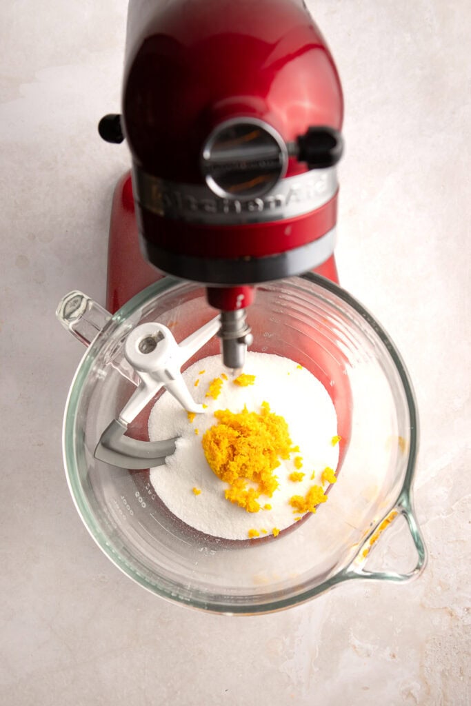 Bright red stand mixer blending sugar and lemon zest in a glass mixing bowl, perfect for baking desserts and sweets.