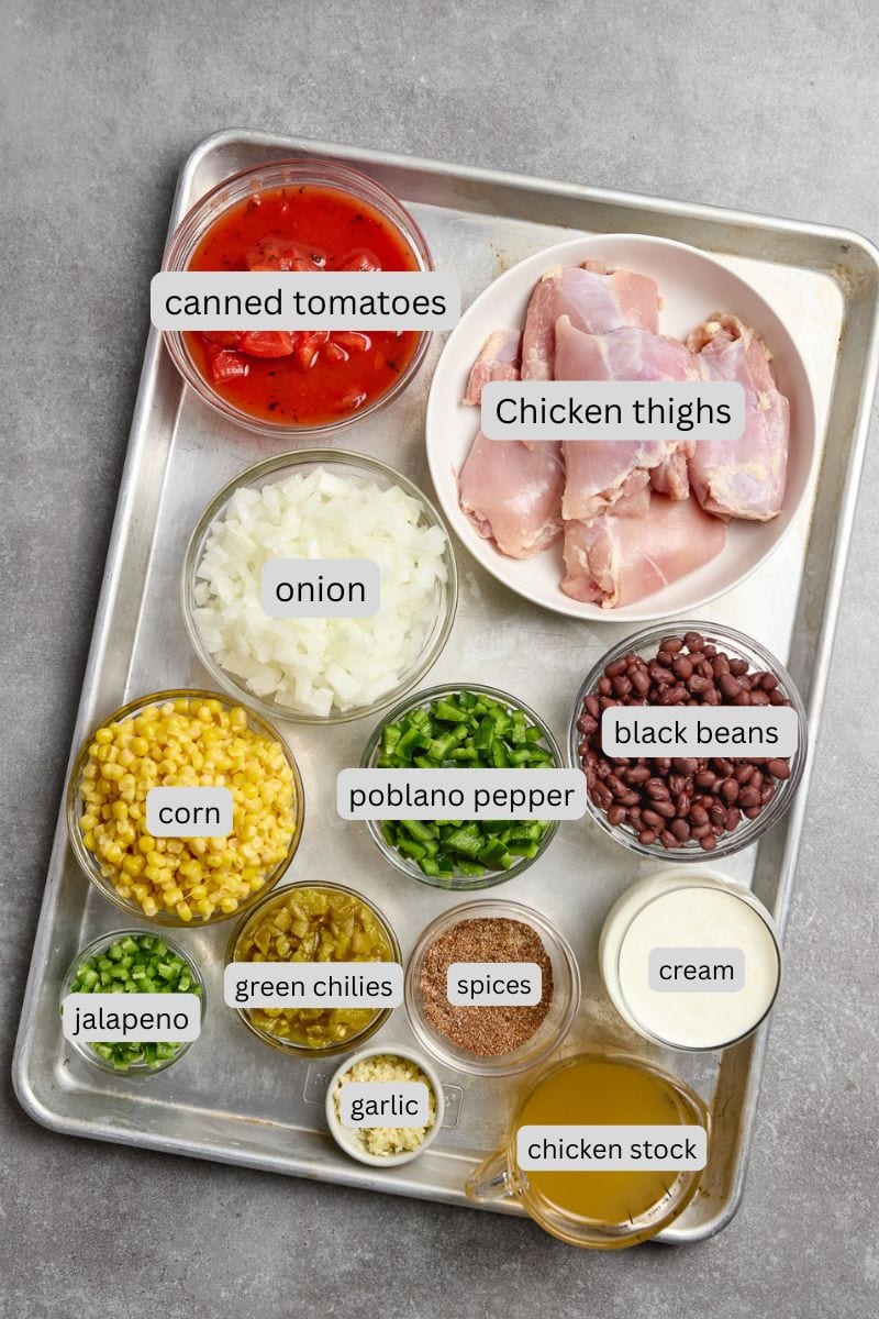 Canned tomatoes, chicken thighs, onion, black beans, corn, poblano pepper, jalapeno, green chilies, spices, garlic, cream, chicken stock on a baking tray for Mexican chicken chili recipe.