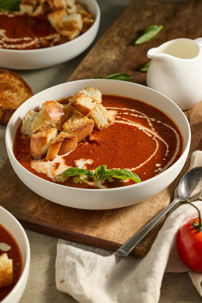 Creamy tomato basil soup with croutons in a white bowl, served with fresh basil and a small pitcher of cream on rustic wooden table, perfect for comfort food lovers.