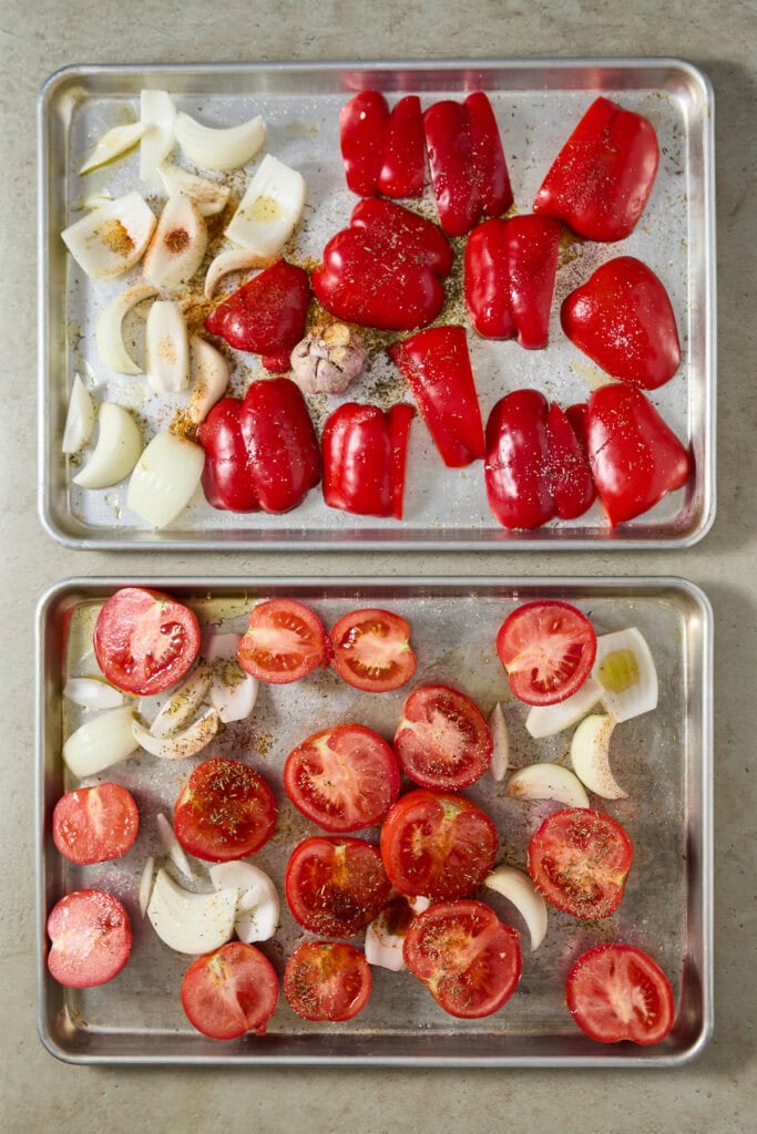 Fresh chopped red bell peppers, tomatoes, and garlic on baking sheets for a recipe at Stuck On Sweet.