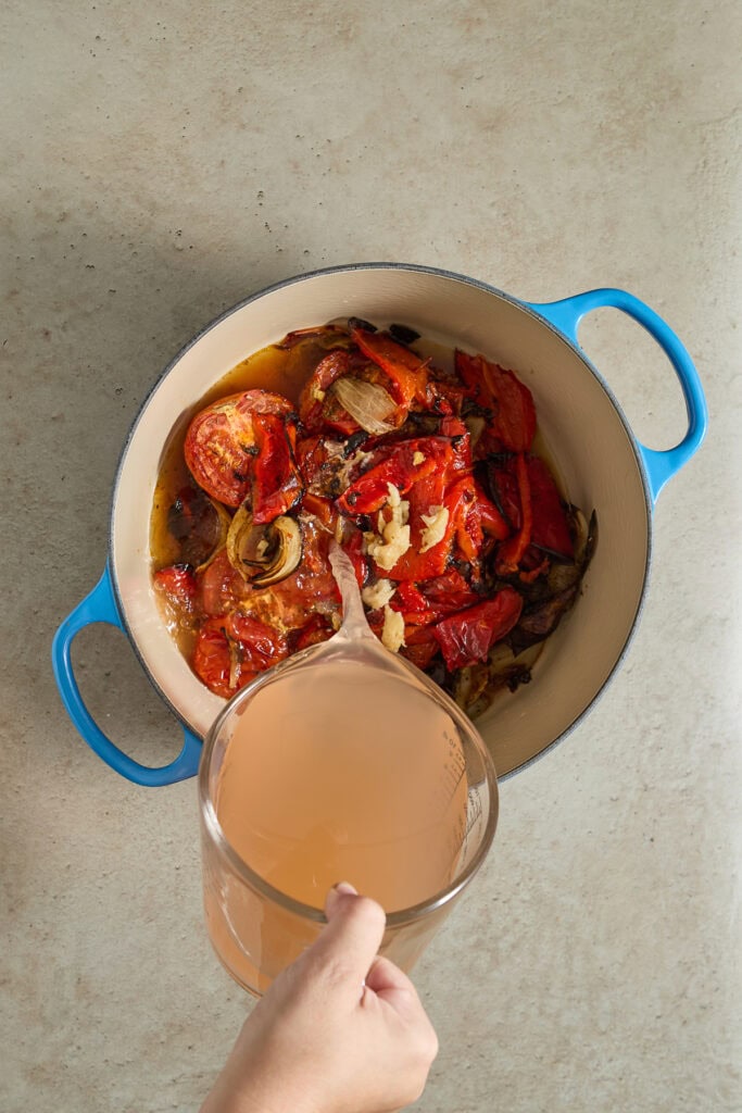 Stew with tomatoes, onions, and garlic in a light-colored pot, with a hand pouring a peach-colored beverage into it.