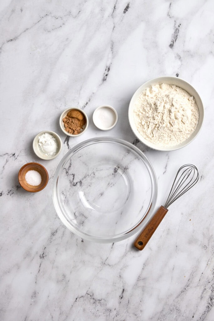 Cream-colored mixing bowl and small bowls with flour, baking soda, salt, cinnamon, and vanilla extract on marble surface for baking.