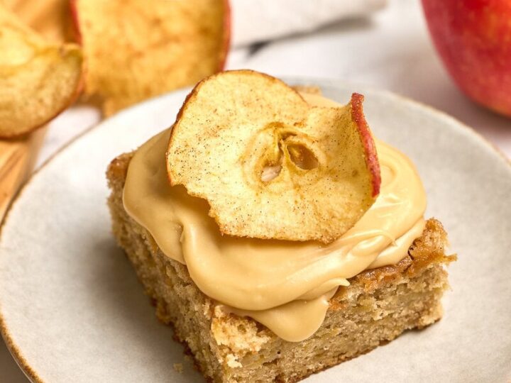 Fresh spiced apple cake square topped with creamy brown sugar frosting and dried apple slices, served on a white plate with a whole apple and apple chips in the background. Perfect for apple dessert lovers.