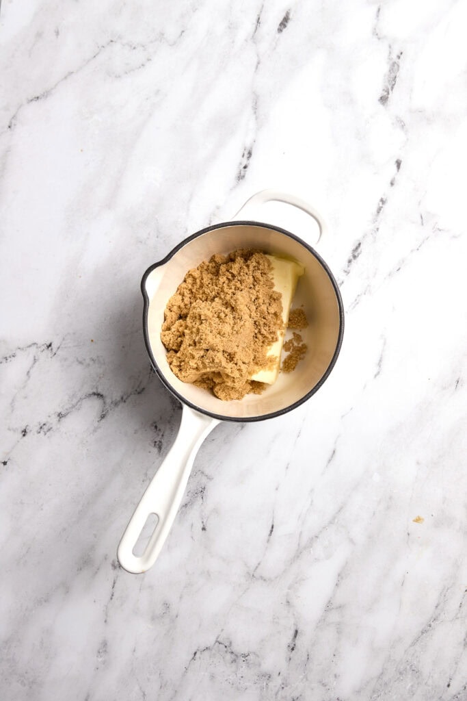 Butter with brown sugar and graham cracker crumbs in a small white saucepan on white marble background.