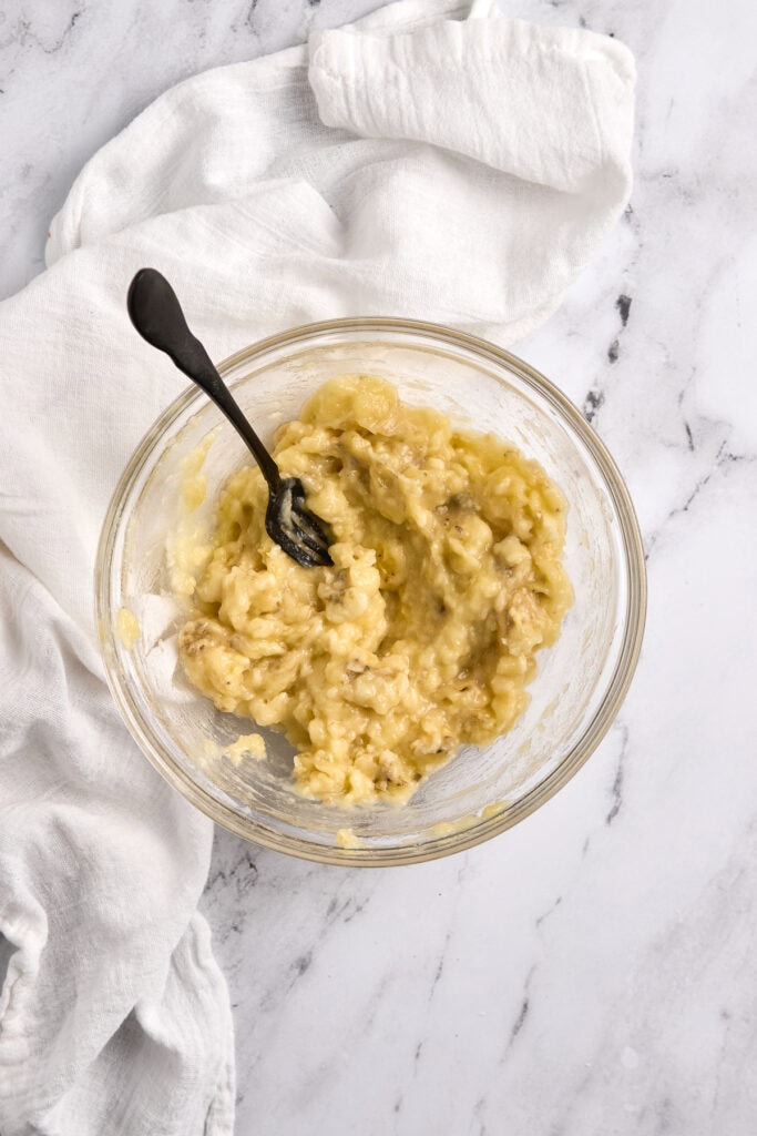 Creamy homemade banana pudding in a glass bowl with a black spoon, on white marble background and white cloth, dessert recipe, comfort food, sweet treat, Stuck On Sweet.