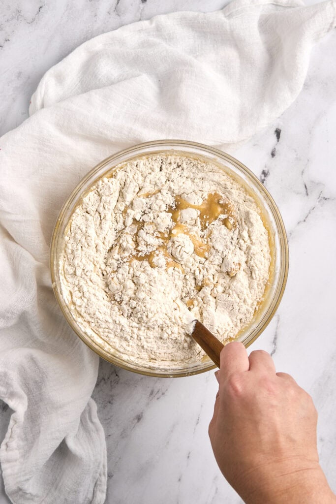 Creamy pie filling being mixed with flour and caramel in a glass bowl, on white marble surface with a white cloth, ready for dessert preparation.
