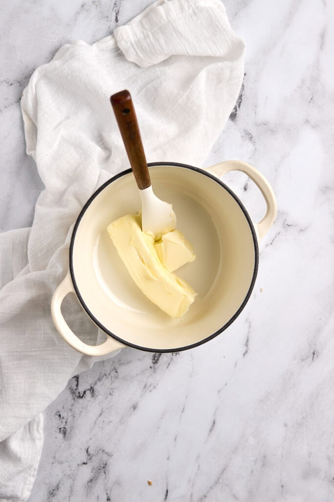 Creamy butter in a white enamel saucepan, ready for cooking or baking, with a wooden-handled spatula, on a marble countertop, ideal for dessert recipes or baking ingredients.