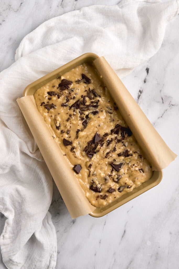 Rich chocolate chip cookie dough ice cream in a beige baking dish with parchment paper edges, topped with chopped dark chocolate, served on a white marble countertop with a white linen cloth.