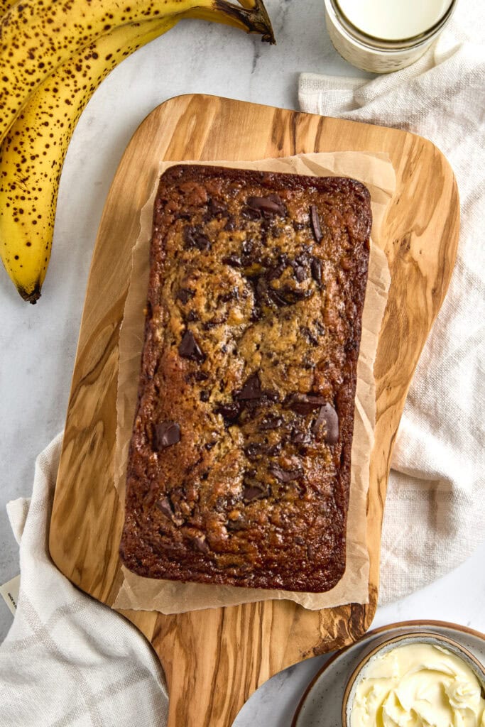 Banana bread with chocolate chunks on wooden cutting board, fresh bananas and glass of milk, perfect for breakfast or snack.