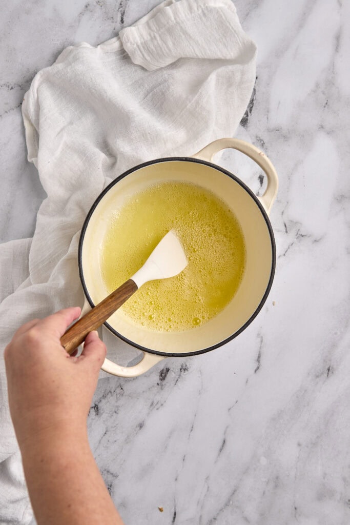 Butter melting in a saucepan with a wooden spatula on a marble countertop with a white kitchen towel.