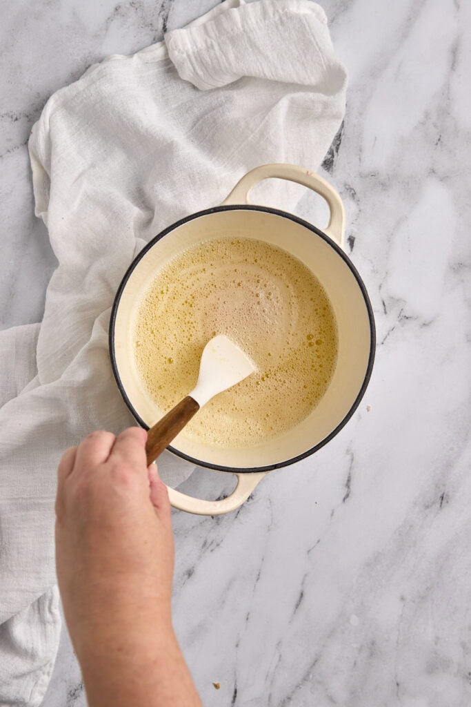 Cream-colored pot with a creamy mixture and a wooden spatula on a marble surface, with white cloth nearby.