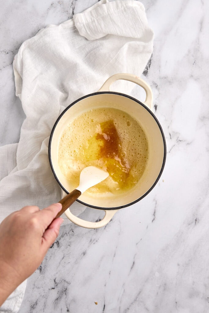 Sweet caramel sauce being prepared in a cream-colored pot on a marble countertop, with a white cloth nearby.