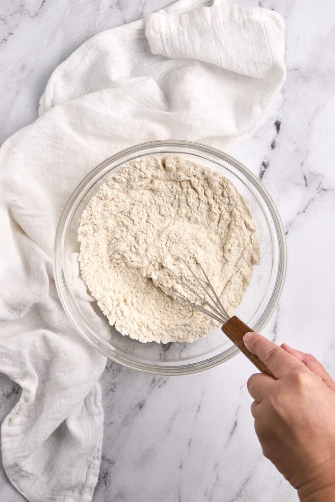 Fine white flour in a glass mixing bowl with a hand whisk, ready for baking, on a white marble surface with a white cloth, ideal for cake and pastry recipes.