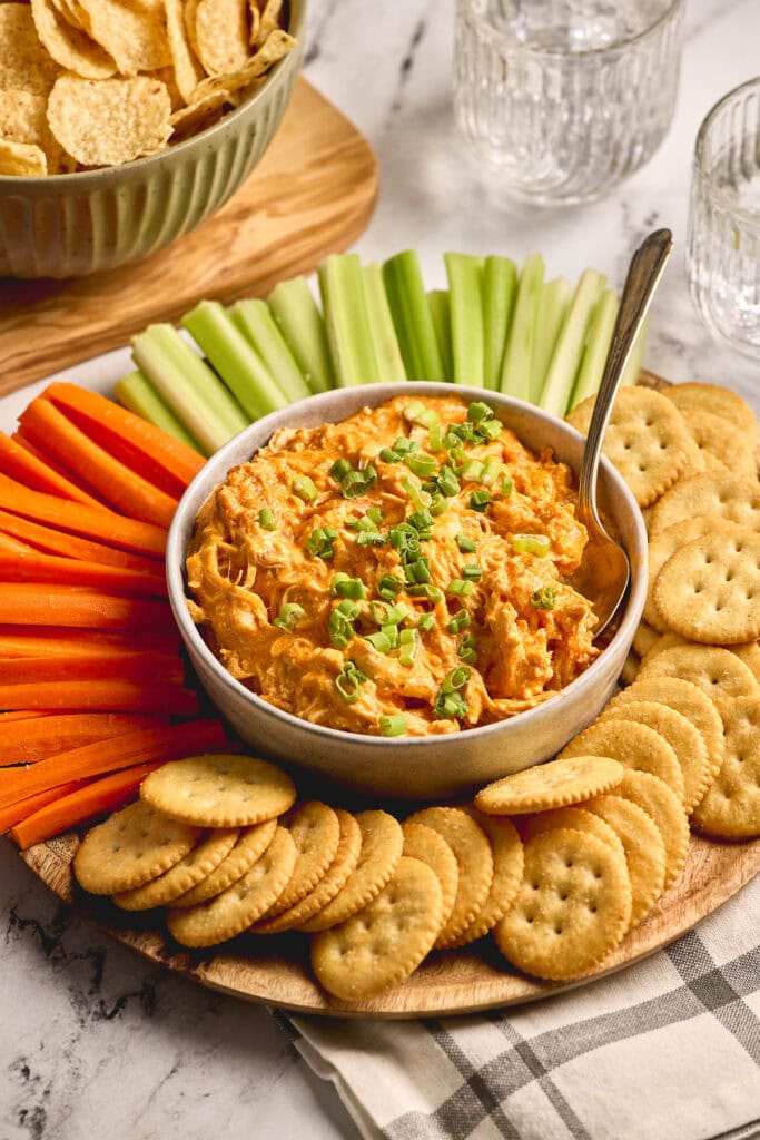 Creamy buffalo chicken dip served with fresh celery sticks, carrot sticks, and buttery crackers on a wooden platter, perfect for game day or parties.
