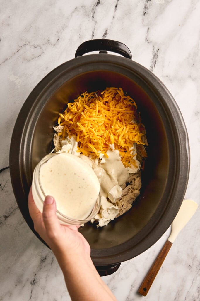Creamy chicken and shredded cheese being added to a slow cooker with cooked chicken and ingredients, preparing a delicious cheesy chicken dish.
