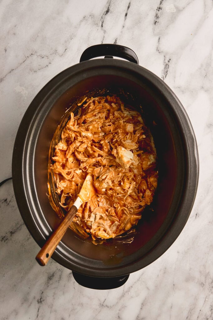Shredded barbecue chicken cooking in a slow cooker with a wooden spatula.