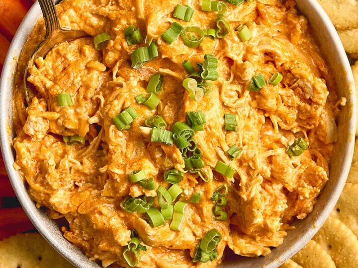 Creamy buffalo chicken dip garnished with chopped green onions and served with saltine crackers and fresh vegetable sticks for a tasty appetizer.