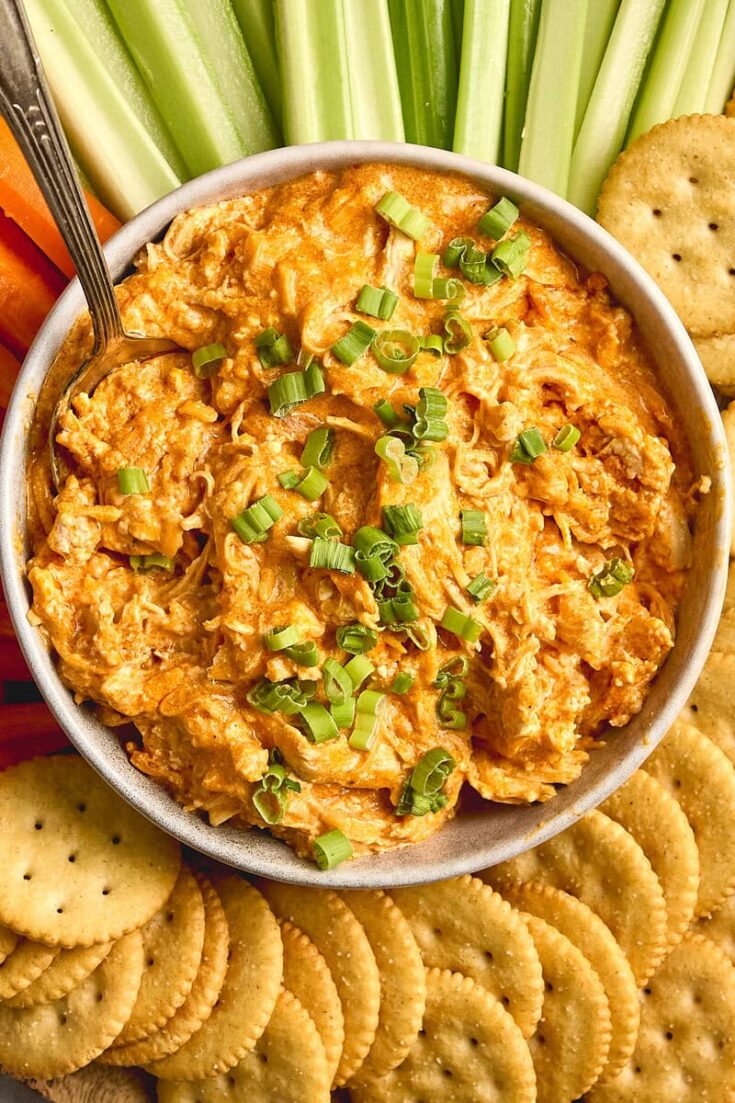 Creamy buffalo chicken dip garnished with chopped green onions and served with saltine crackers and fresh vegetable sticks for a tasty appetizer.