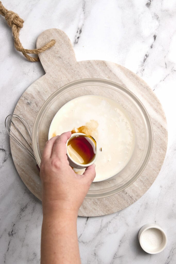 Creamy milk mixture with vanilla extract in glass bowl for dessert recipe, on marble surface.