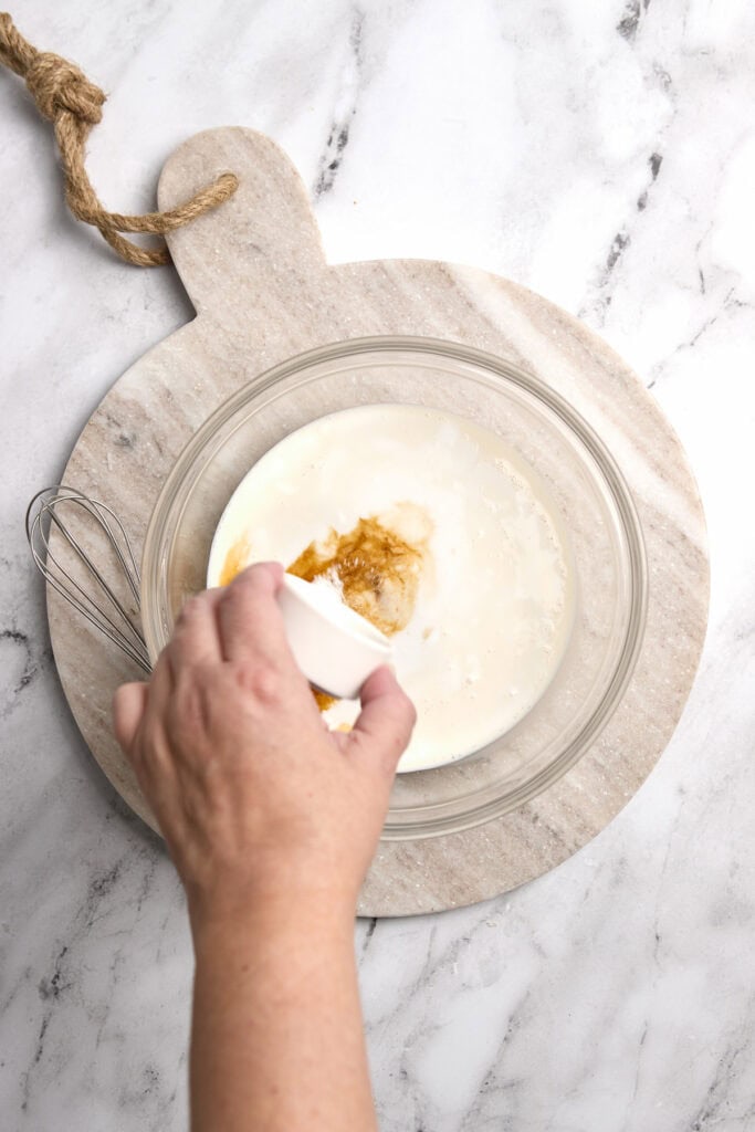 Cream being poured into a mixing bowl with sugar on a marble countertop, preparing a sweet dessert.