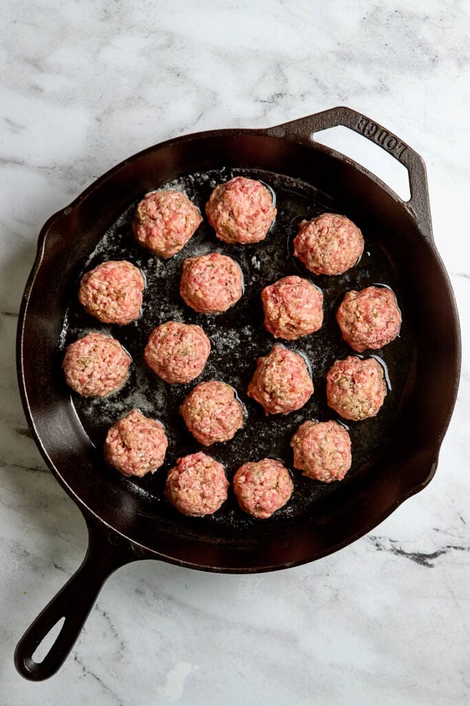 Homemade meatballs cooking in a cast iron skillet on a white marble countertop, ready to be served as an appetizer or meal.