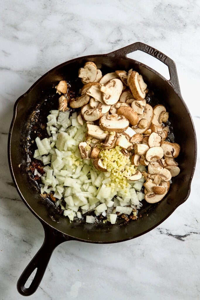 Sautéed chopped onions, garlic, and sliced mushrooms cooking in a cast iron skillet for a savory recipe. Healthy ingredients prepared for an easy, delicious meal or side dish.