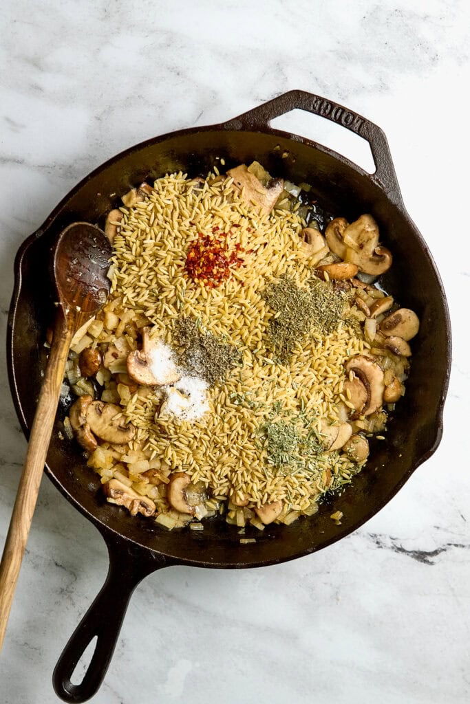 Sautéed mushroom and onion skillet with dry pasta, seasoned with herbs and spices, in cast-iron pan.