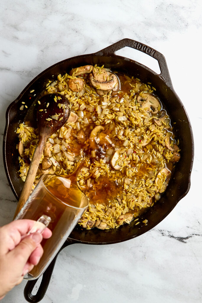 Creamy mushroom and rice skillet dish with broth being poured in.