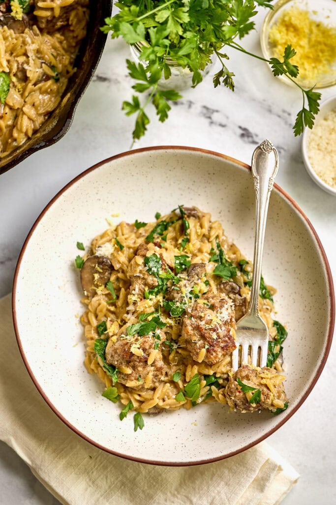 Creamy mushroom orzo with spinach and meatballs garnished with fresh parsley, served in a white bowl with grated cheese and herbs, perfect comfort food for dinner or lunch.