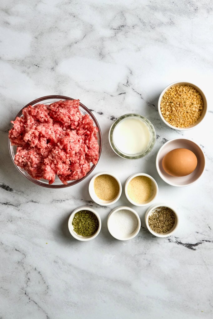 Freshly prepared ground meat and ingredients for meatballs on marble countertop, including breadcrumbs, egg, milk, herbs, and seasonings, ready for baking or cooking.