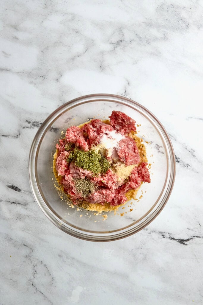 Minced raw meat mixture with herbs and seasonings in a glass bowl, ready for cooking or preparing a dish, on a white marble surface.