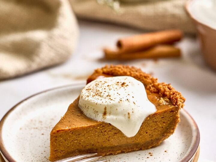 Creamy pumpkin pie with sweetened condensed milk in a graham cracker crust topped with whipped cream and sprinkled with cinnamon, served on a rustic plate with cinnamon sticks and festive spices in the background.