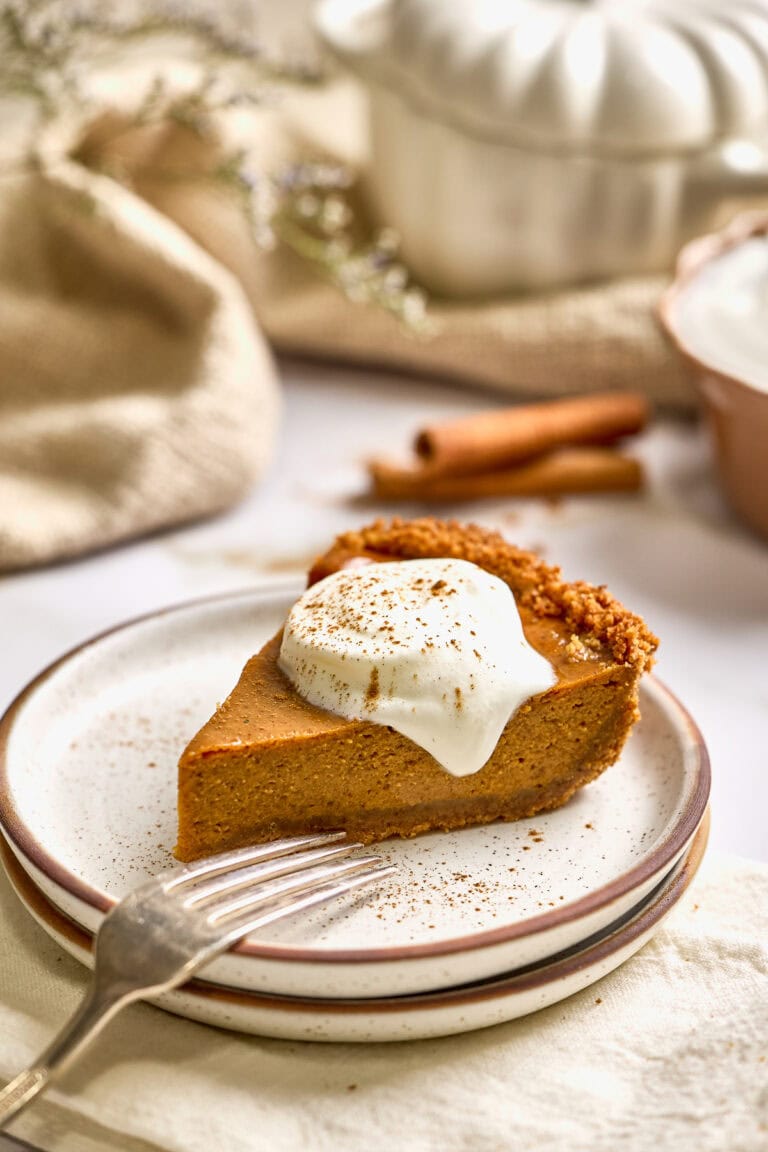 Creamy pumpkin pie with sweetened condensed milk in a graham cracker crust topped with whipped cream and sprinkled with cinnamon, served on a rustic plate with cinnamon sticks and festive spices in the background.