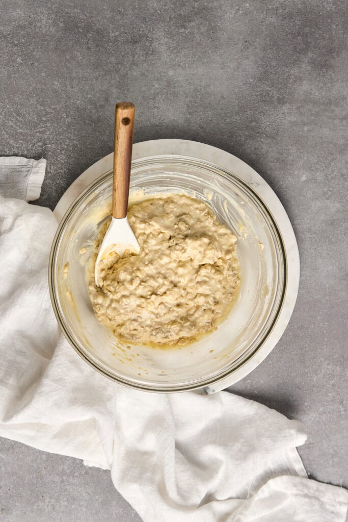 Creamy oatmeal muffin batter in glass mixing bowl, ready for baking, on a gray textured surface with white cloth accent. Perfect for banana bread and muffin recipes.