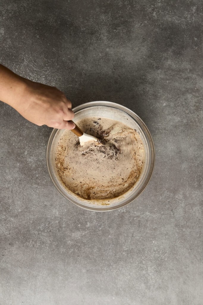 Hand stirring creamy chocolate soft serve ice cream in a glass bowl with a spatula, ready for dessert, highlighting homemade ice cream recipes.