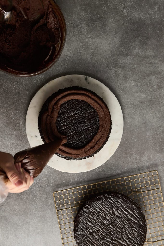 Rich chocolate cake being decorated with piping bag of chocolate frosting, on marble cake stand, with cooling rack and extra cake layer nearby, perfect for dessert or special occasions.