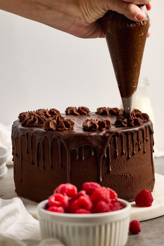 Rich chocolate cake being decorated with luscious fudge ganache from a piping bag, surrounded by fresh raspberries in a white bowl for a delicious dessert presentation.