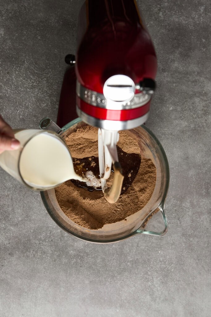 Close-up of hand pouring milk into a chocolate cake batter in a mixing bowl with a stand mixer, perfect for cake baking or dessert recipes.