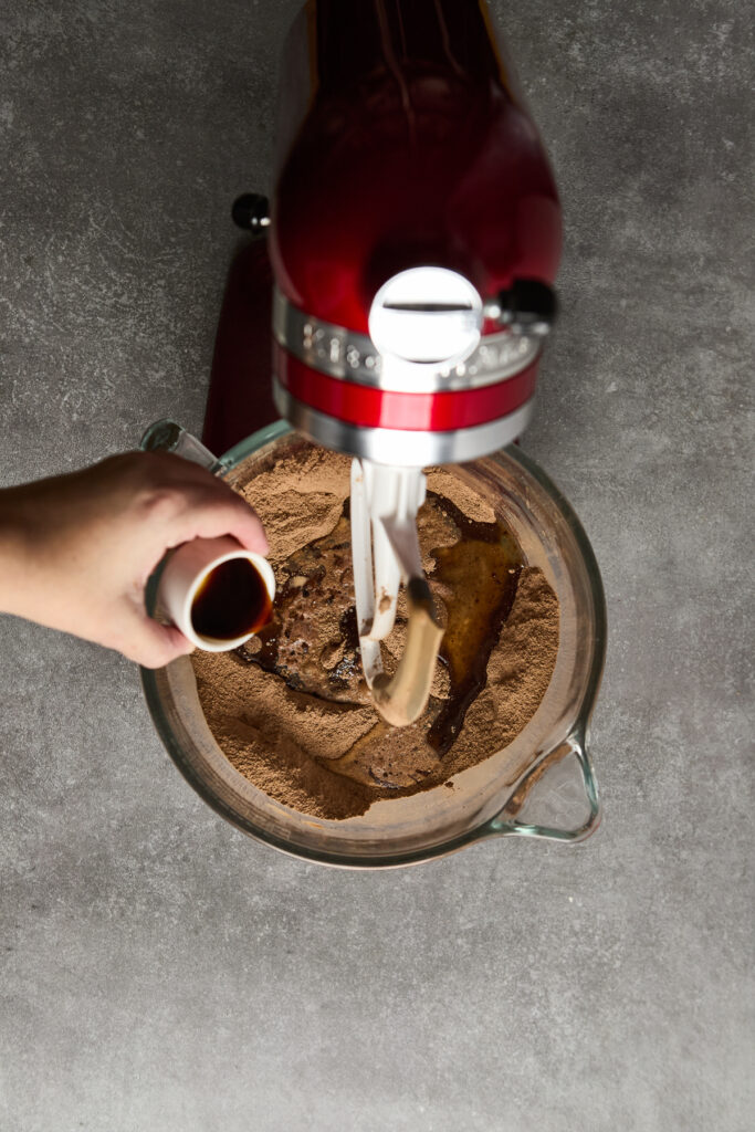 Hand pouring vanilla extract into chocolate cake mix in mixing bowl, with stand mixer on gray countertop.