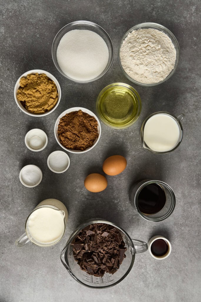 Delicious ingredients for making rich chocolate ganache cake, including eggs, sugar, flour, cocoa, butter, and chocolate, arranged on a gray surface perfect for baking or dessert recipes.