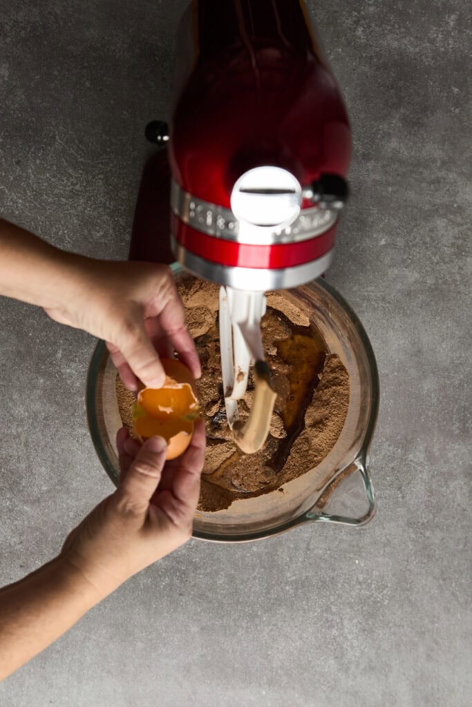 Hand cracking eggs into mixing bowl for chocolate cake batter, with stand mixer in the background.