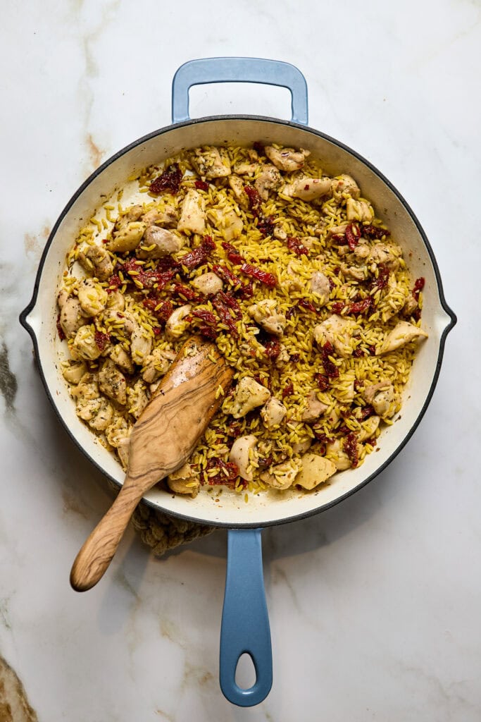 Delicious chicken and rice skillet dish with sun-dried tomatoes, herbs, and tender cooked chicken in a white enamel pan with a blue handle. Perfect for easy weeknight meals or comfort food.